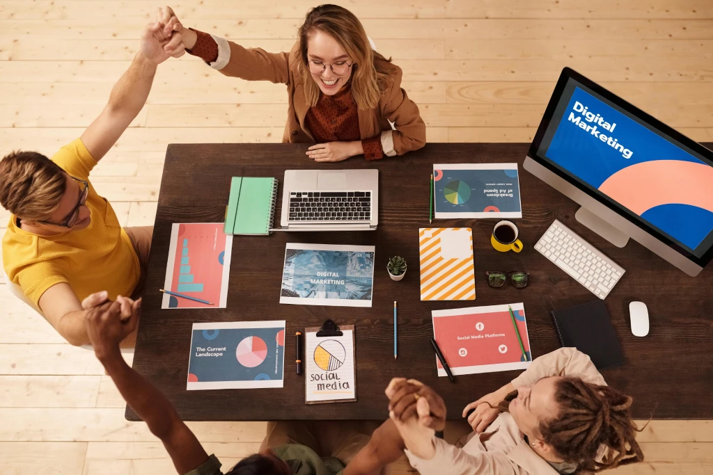 Four people sit at a table with laptops, notebooks, and digital marketing papers, holding hands in a celebratory gesture. A computer screen on the table displays "Digital Marketing.