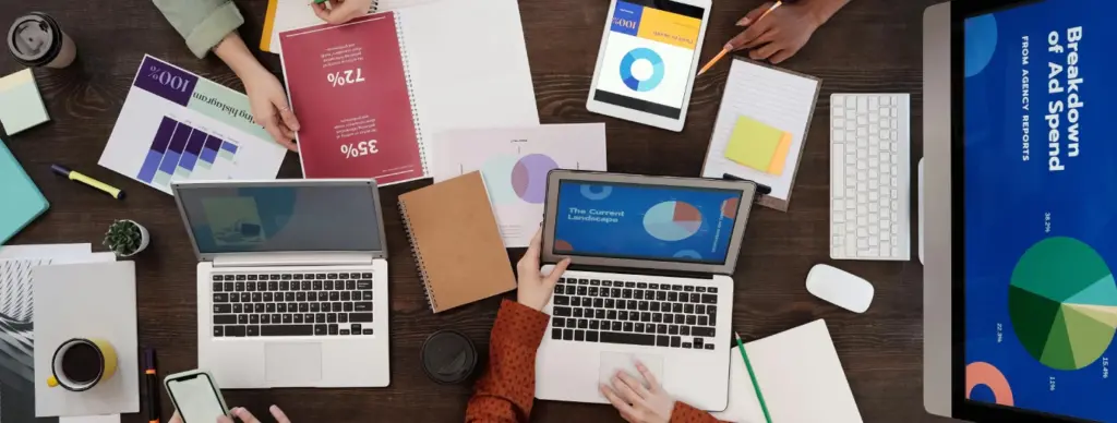 A group of people working at a table with laptops, tablets, documents, and charts, collaborating on a project.
