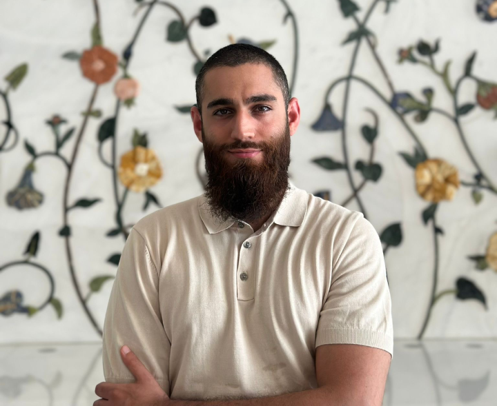 A man with a beard and a short-sleeved collared shirt stands in front of a floral wall, embodying the essence of Small World Marketing's About Us ethos.