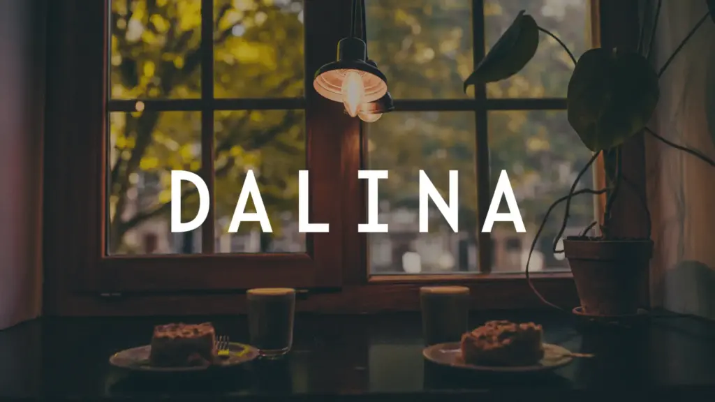 Two plates with cake and two glasses sit on a table by a window, with a hanging lamp and a potted plant. The word DALINA is centered over the image. Trees are visible outside the window.
