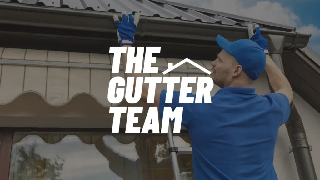 A worker in blue uniform stands on a ladder, cleaning gutters on a house. The text “THE GUTTER TEAM” with a roof icon appears over the image.