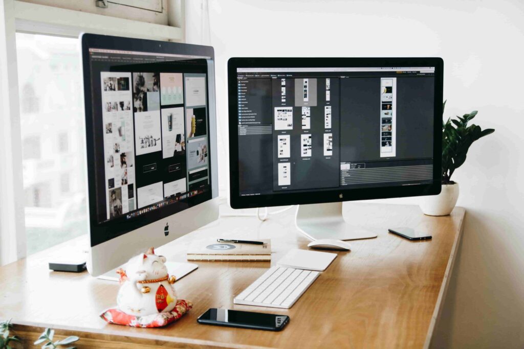 Two large computer monitors on a wooden desk display design software, inspiring 2026 Best Website Design Ideas for Immigration Law Firms. The workspace features a keyboard, mouse, notebooks, smartphone, potted plant, and ceramic cat figurine with red scarf.