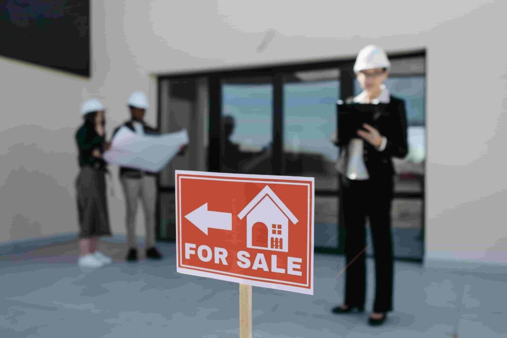 A For Sale sign with a house icon is in focus, while three people in hard hats—reflecting 2026 Best Website Design Ideas for Real Estate Agencies—examine blueprints and a clipboard outside a building in the background.