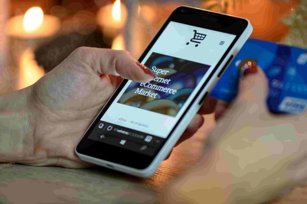 A person holds a smartphone displaying an online shopping website from https://smallworld.ca/website-design/ in one hand and a blue credit card in the other, suggesting they are making a purchase. Warm lights are blurred in the background.