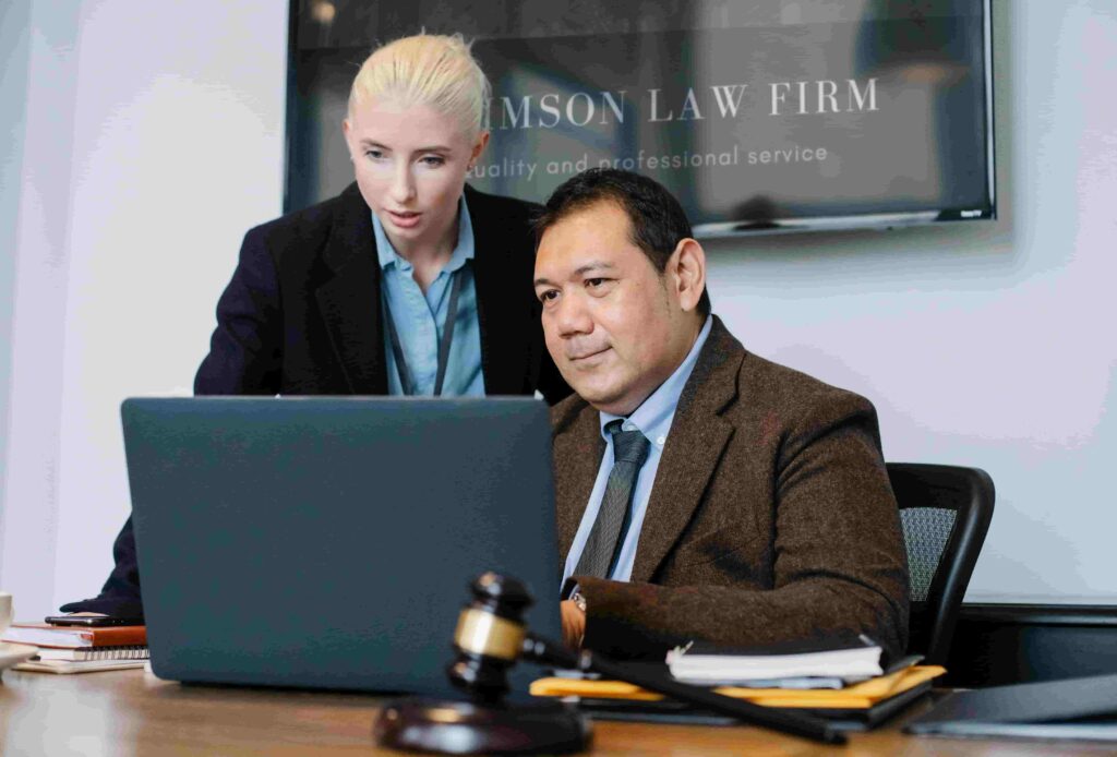 Two lawyers in formal attire look at a laptop in an office. A gavel, documents, and a pen are on the desk. A sign reading LAW FIRM is visible, suggesting they’re reviewing the Best 9 Ways to Generate Leads for Employment Law Firms.
