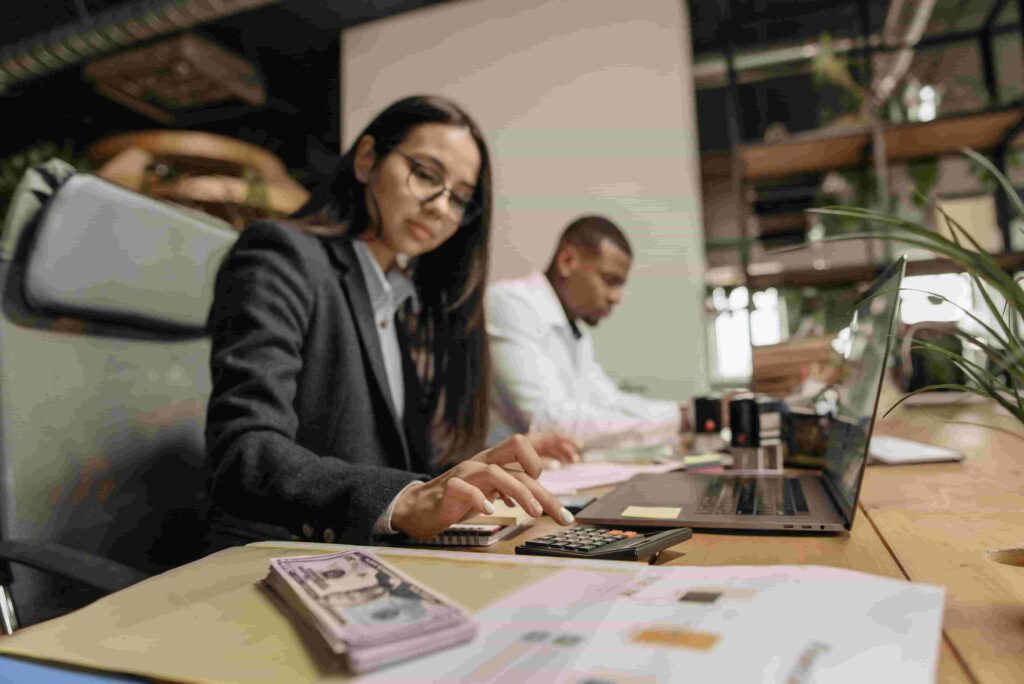 A woman in business attire uses a calculator at a desk with cash and documents, while a man works on a laptop in the background—an office scene reflecting how accounting firms boost SEO and attract more clients.