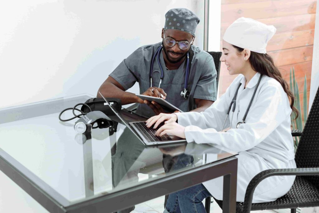 Two healthcare professionals sit at a glass table, one working on a laptop and the other using a tablet, possibly researching How Can Healthcare Providers Use SEO for New Patients? Both wear medical scrubs and collaborate in a bright, modern office.