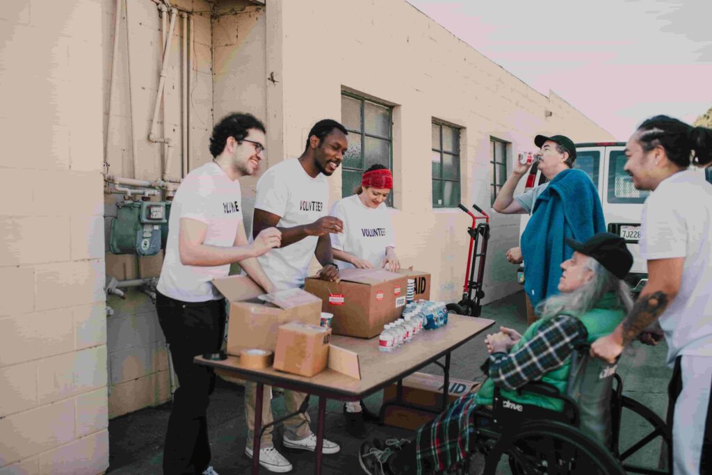 A diverse group of volunteers stands around a table with food and drinks, smiling and interacting with an older woman in a wheelchair outside a building—showing how nonprofits can use content marketing to increase donations through community engagement.
