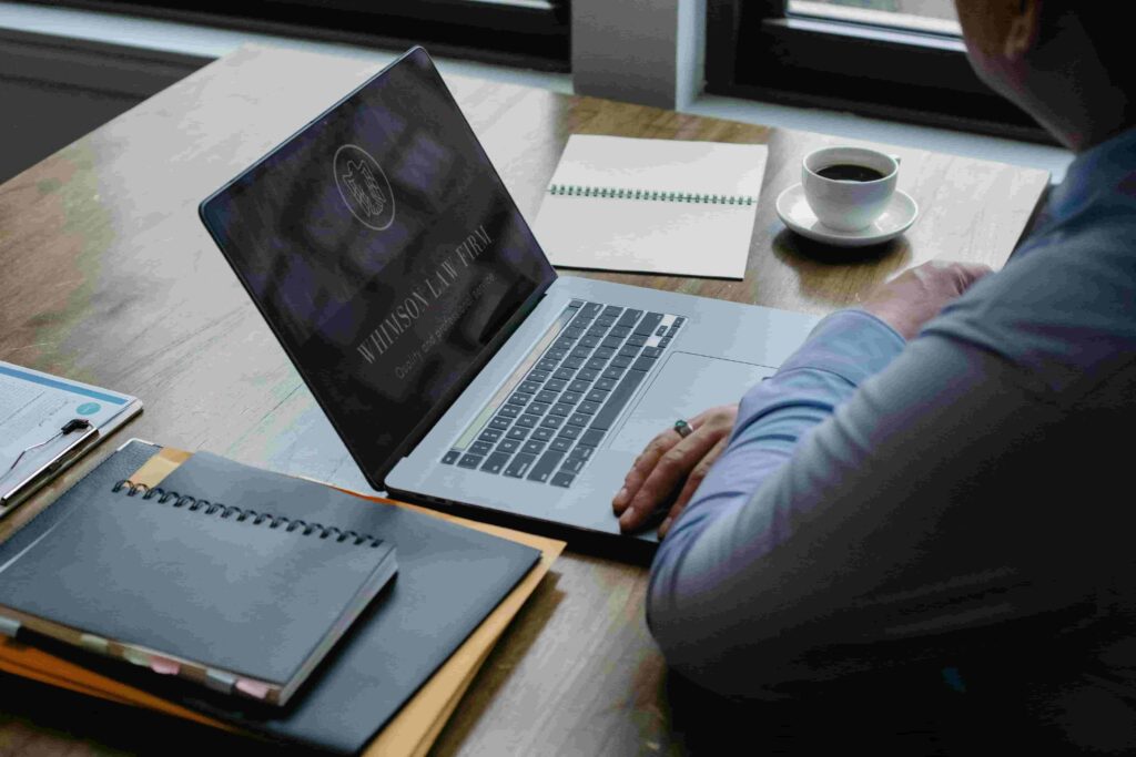 A person sits at a wooden desk using a laptop, researching How to Generate Leads for Immigration Law Firms. Notebooks, a notepad, and coffee sit nearby as the screen displays the law firm logo and “Winston Law Firm.”.