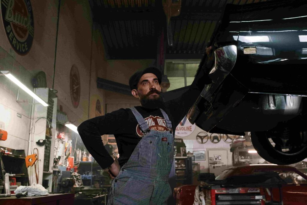 A mechanic with a beard and overalls stands in a dimly lit garage, looking at a car raised on a lift. The workshop, cluttered with tools and signs, hints at top marketing strategies for auto repair shops to generate more leads.
