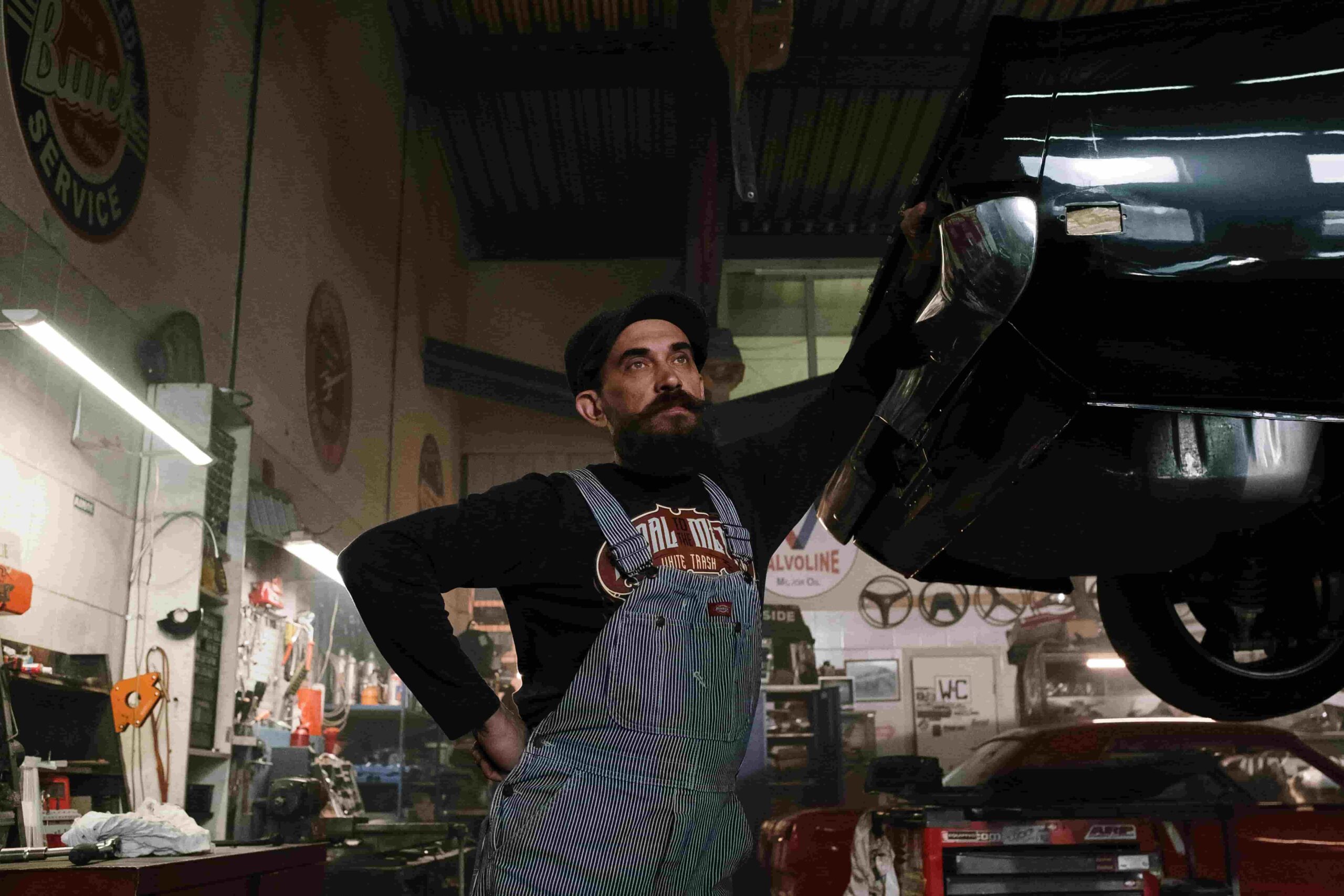 A mechanic with a beard and overalls stands in a dimly lit garage, looking at a car raised on a lift. The workshop, cluttered with tools and signs, hints at top marketing strategies for auto repair shops to generate more leads.